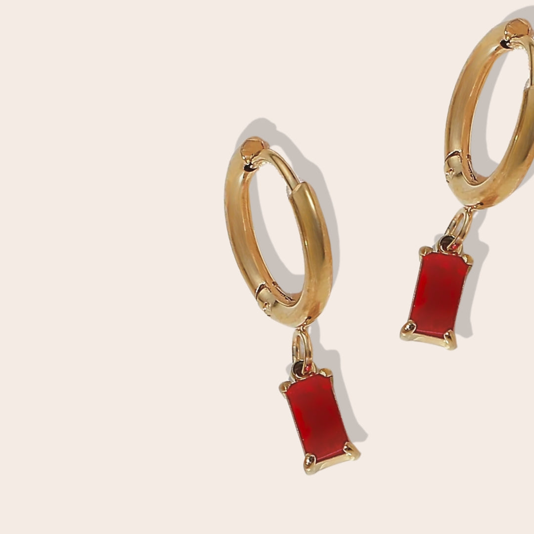 Close up of Rhea Rectangular Huggies in ruby. Made from stainless steel and plated with 18k gold, these earrings are inlaid with sparkling cubic zirconia.