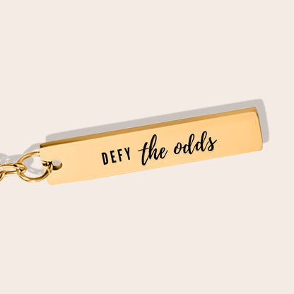 Vertical Bar Keychain - Defy the Odds in gold close up. Crafted from stainless steel and plated with 18K gold.