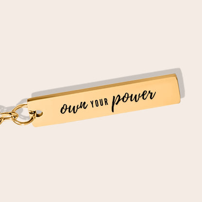 Vertical Bar Keychain - Own Your Power in gold, close up. Crafted from stainless steel and adorned with lustrous 18K gold plating, this chic piece features a laser-engraved affirmation.