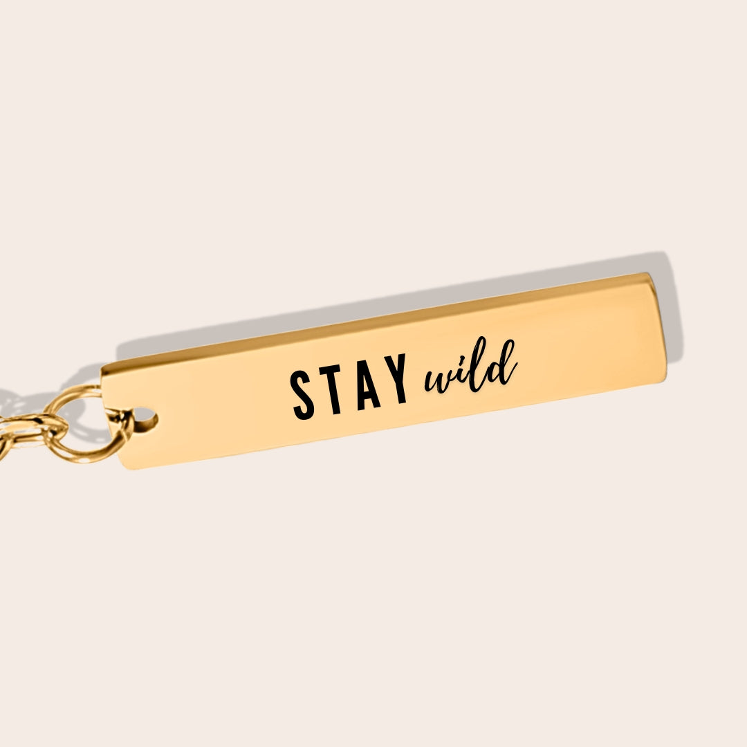 Vertical Bar Keychain - Stay Wild, in gold, close up. Made from durable stainless steel and adorned with 18K gold plating, this sleek piece features a laser-engraved reminder.